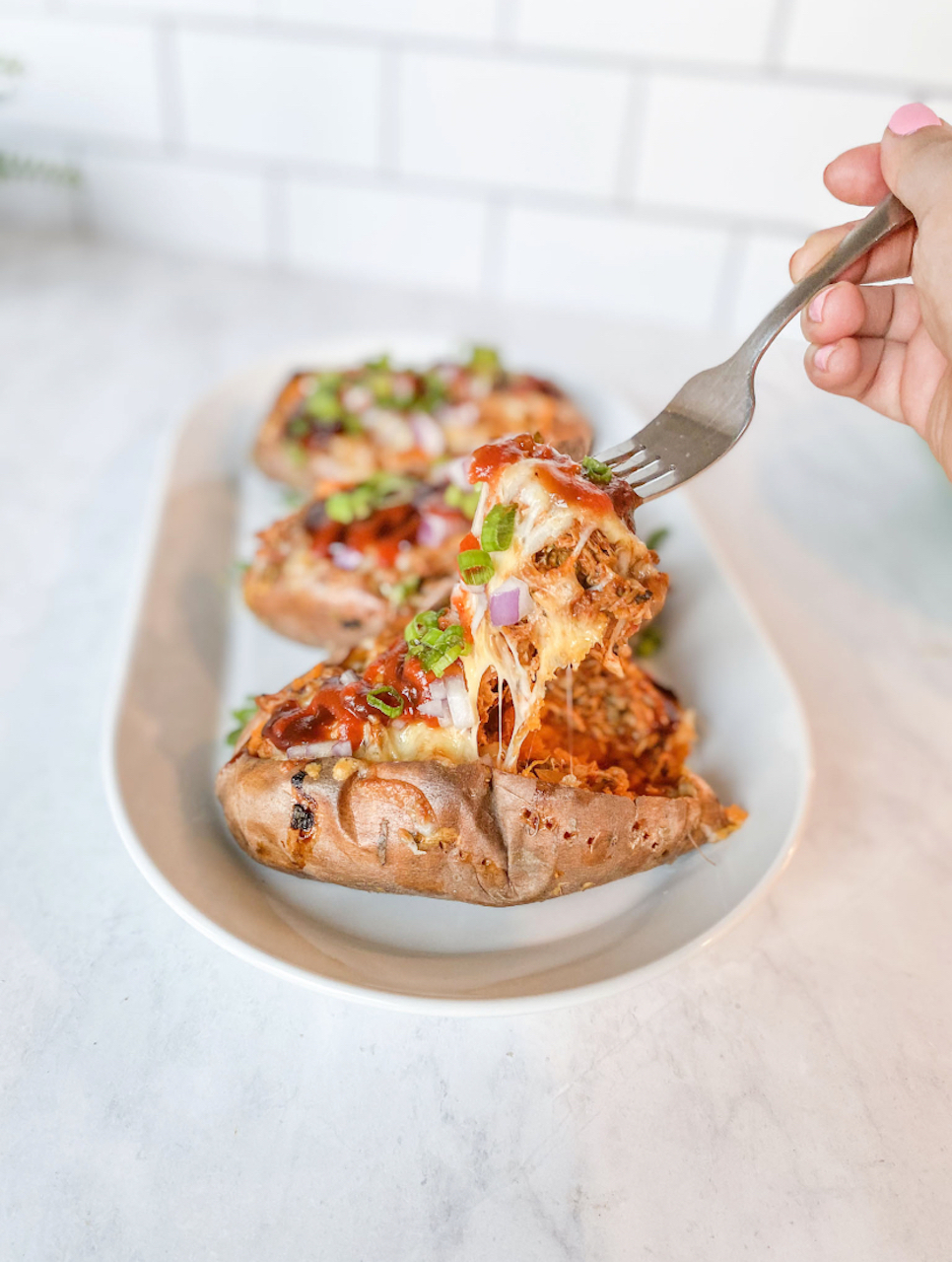 BBQ Chicken Stuffed Sweet Potato recipe image with three stuffed sweet potatoes filled with shredded BBQ chicken topped with cheese, red onions and green onions. Metal fork is performing a cheese pull with recipe ingredients.  