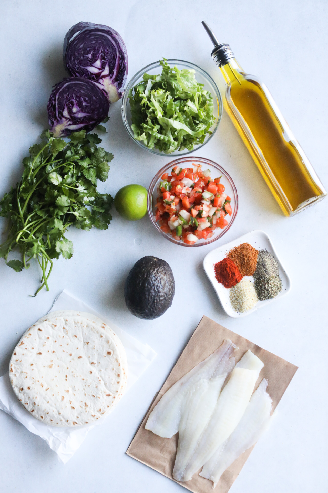 Blackened Fish Tacos ingredients on a flat lay. Olive oil bottle, shredded lettuce, pico de gallo, catfish, cilantro, purple cabbage, tortillas and blackening seasoning.