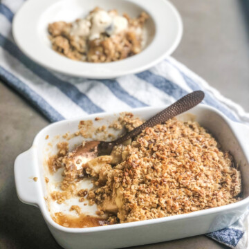 Apple Crumble in baked dish, scooped out and plated on a bowl behind the baked dish that's out of focus. Blue and white striped towel underneath the dishes to add style.