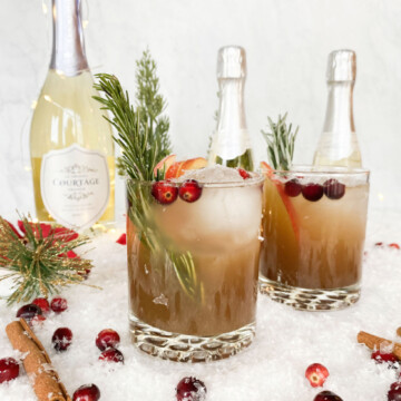 two cocktails in a shakers glass with big round ball of ice mold with apple cider bourbon cocktail. behind glasses is a bottle of brut with two mini bottles to the left of the bottle. garnishes are fresh cranberry, fake snow and fresh rosemary.