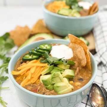 Creamy Chicken Tortilla Soup with two bowls only one bowl in focus of soup in focus with cheddar cheese, avocado, jalapeno, sour cream and tortilla chips as toppings.