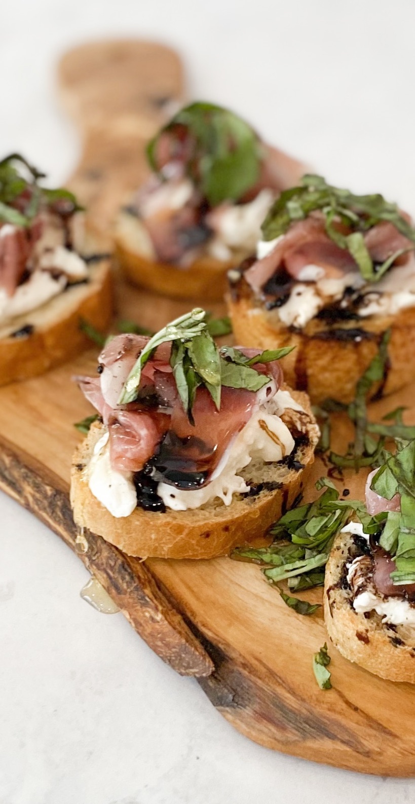 Five prosciutto crostinis in image with one in clear focus. 5 mini crostinis with baked toast, burrata, prosciutto, basil, honey and balsamic drizzle.