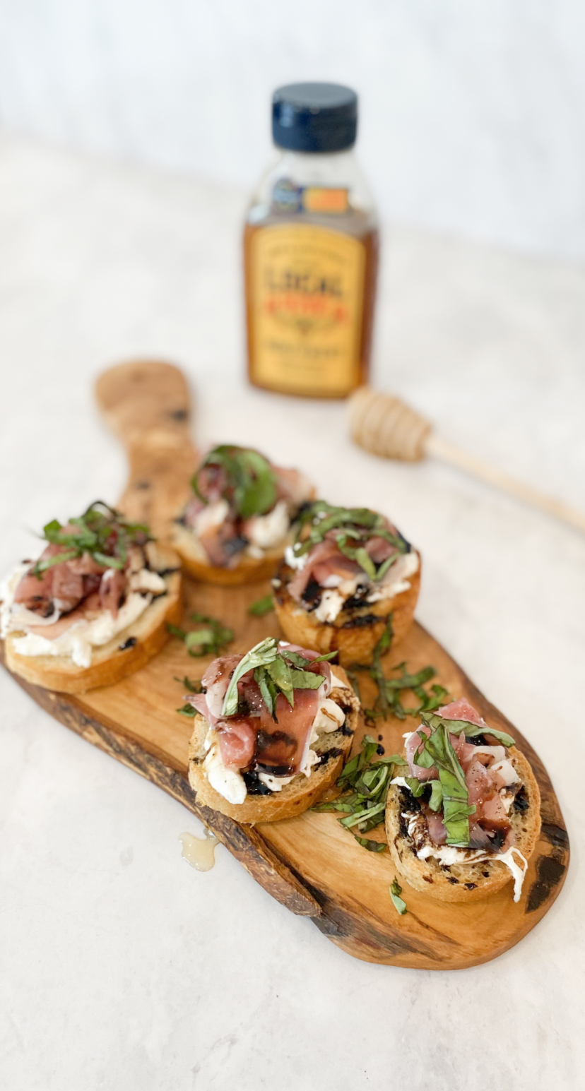 Five prosciutto crostinis in image with one in clear focus. 5 mini crostinis with baked toast, burrata, prosciutto, basil, honey and balsamic drizzle. Local Hive Honey bottle behind crostinis out of focus..