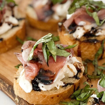 Four prosciutto crostinis in image with one in clear focus. Mini crostinis with baked toast, burrata, prosciutto, basil, honey and balsamic drizzle.