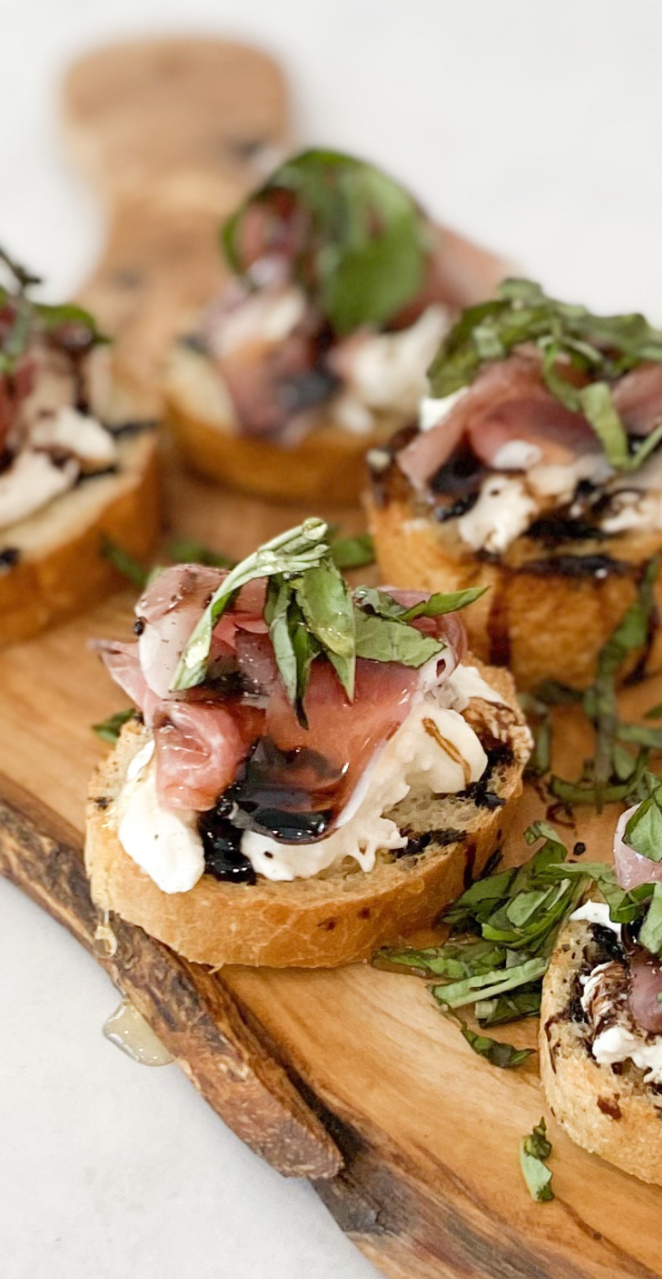 Four prosciutto crostinis in image with one in clear focus. Mini crostinis with baked toast, burrata, prosciutto, basil, honey and balsamic drizzle.