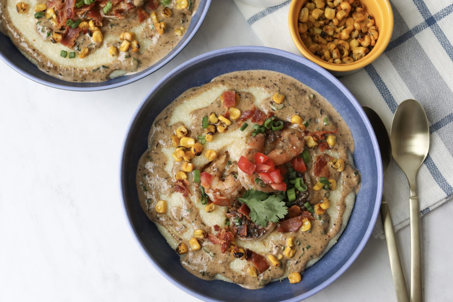 1.5 bowls with easy dirty jerk shrimp and grits. Small bowl of extra roasted corn to the right of the bowl. The jerk shrimp and grits is topped with a cream sauce, jerk shrimp, corn, green onion and cilantro