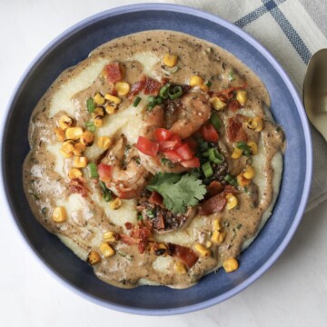 One blue bowl with jerk shrimp and grits. Topped with shrimp, corn, crispy bacon, green onion and cilantro.Bowl is placed on top of a decorative blue plaid towel that matches the bowl and a gold decorative spoon on the right of the bowl.