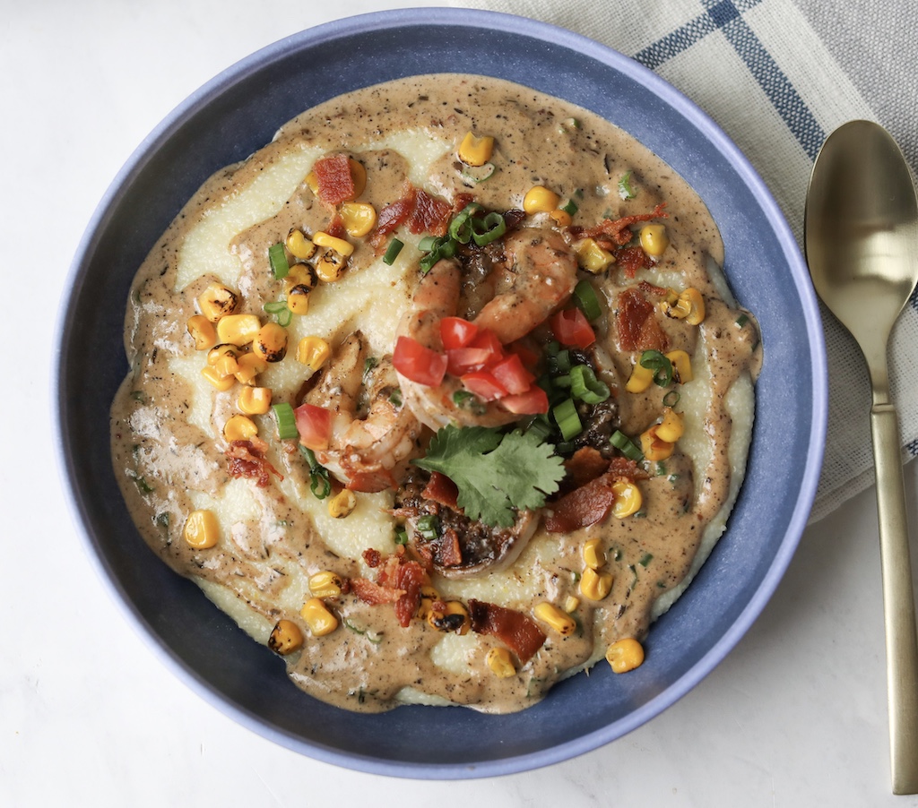 One blue bowl with jerk shrimp and grits. Topped with shrimp, corn, crispy bacon, green onion and cilantro.Bowl is placed on top of a decorative blue plaid towel that matches the bowl and a gold decorative spoon on the right of the bowl.