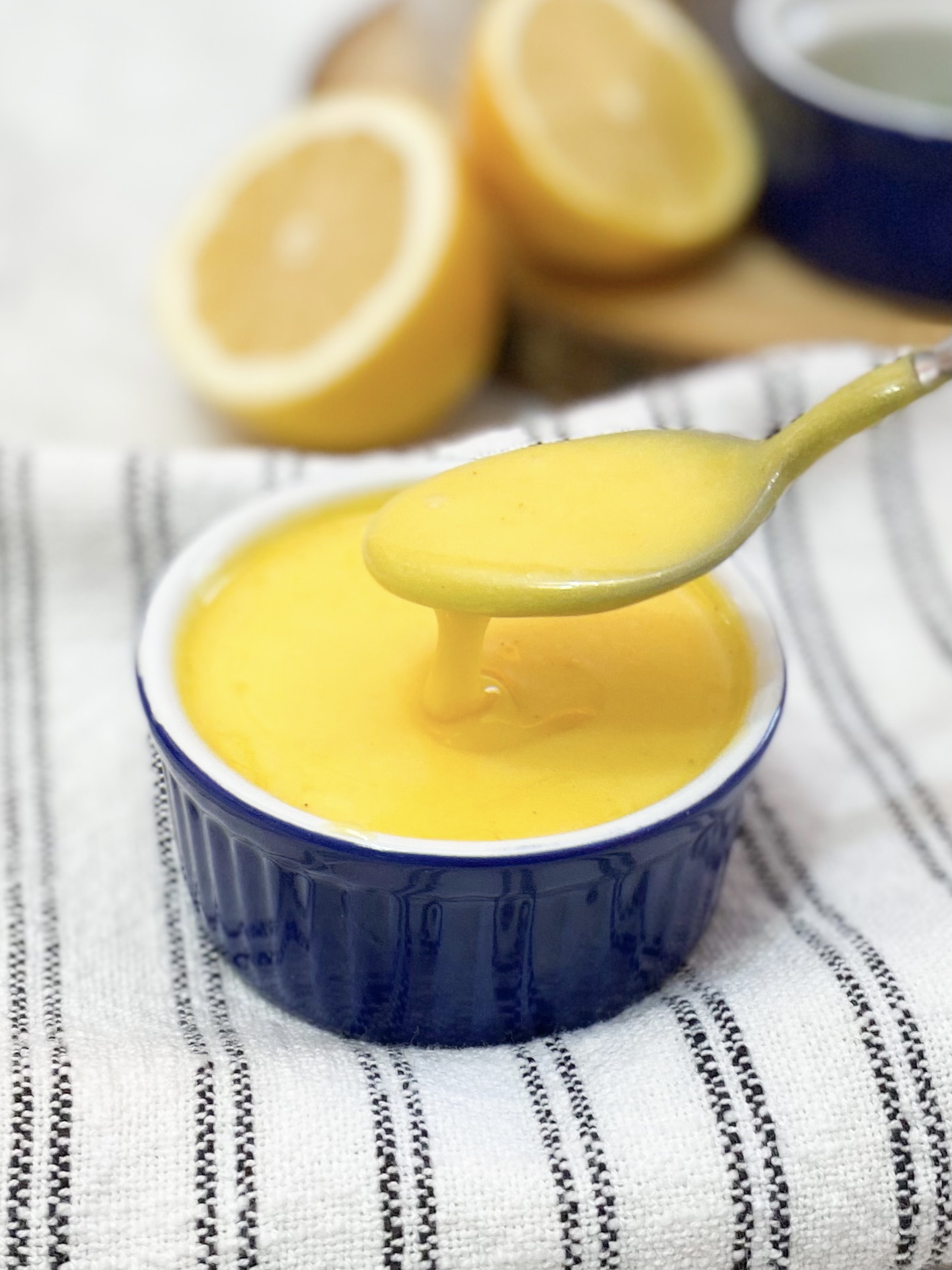 Blue bowl with easy from scratch hollandaise sauce. Spoon is scooping out a portion of the sauce. Blue bowl is sitting on top of black and white striped towel with lemons out of focus in the background.