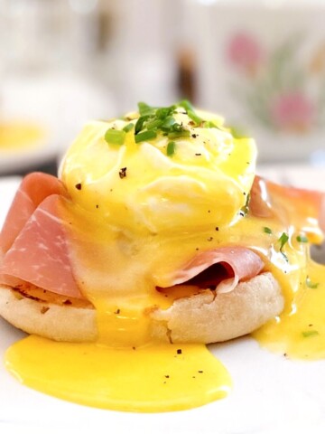 Hollandaise sauce poured over an english muffin with prosciutto, poached eggs and chives.