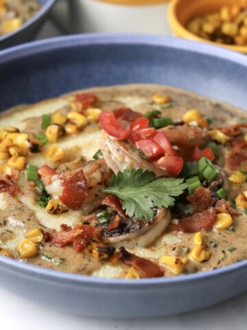 Jerk shrimp and grits plated in a blue bowl topped with crunchy bacon, corn, tomatoes and parsley.