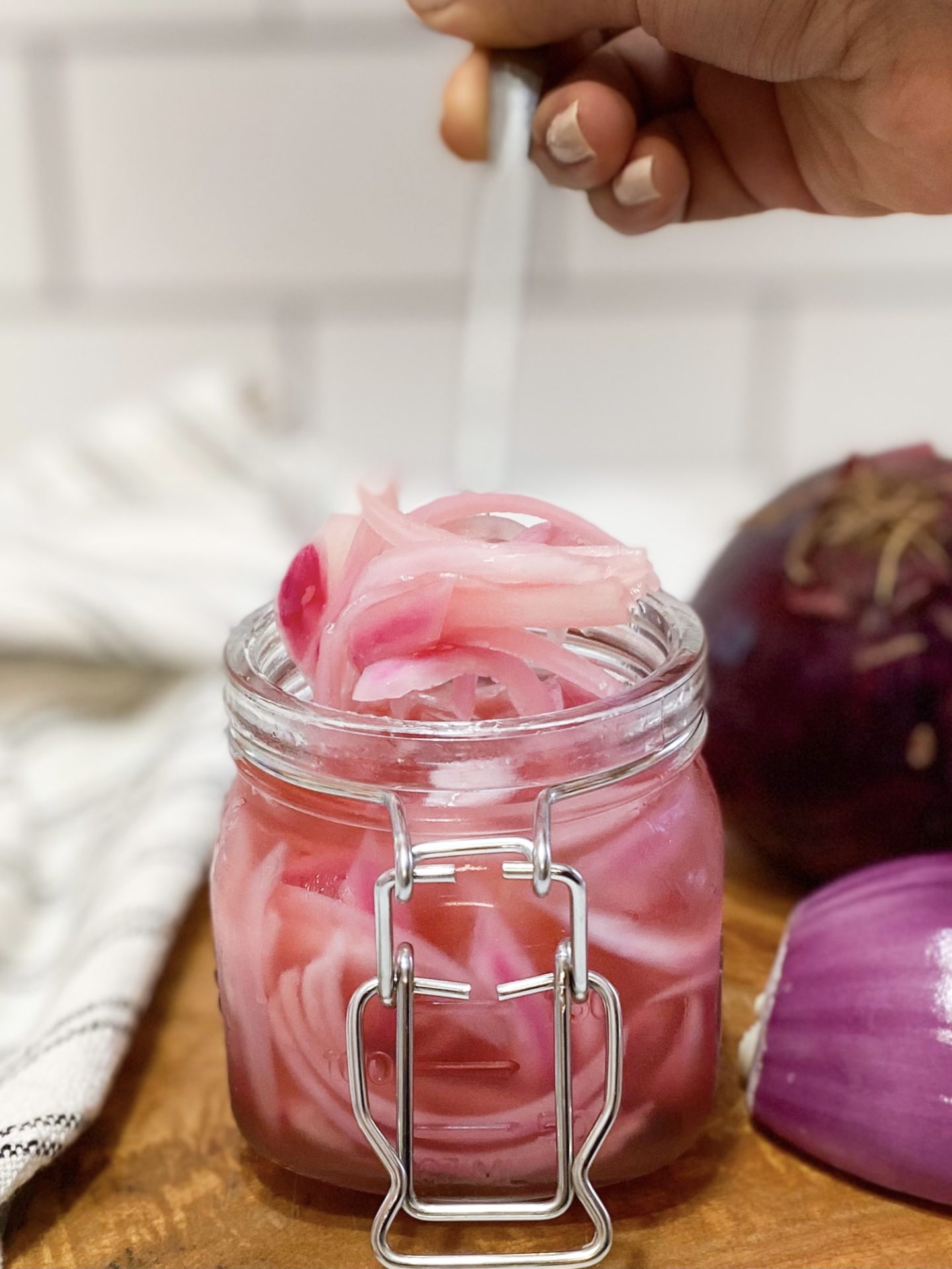 Pickled Red Onions in a small mason jar with latched lid. Jar is sitting on top of a brown small butting board with a sliced onion in half for aesthetic purposes. Hand is present holding a fork, lifting some of the pickled red onions out of the jar to show the finished product.