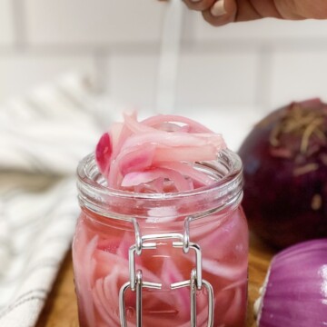 Pickled Red Onions in a small mason jar with latched lid. Jar is sitting on top of a brown small butting board with a sliced onion in half for aesthetic purposes. Hand is present holding a fork, lifting some of the pickled red onions out of the jar to show the finished product.