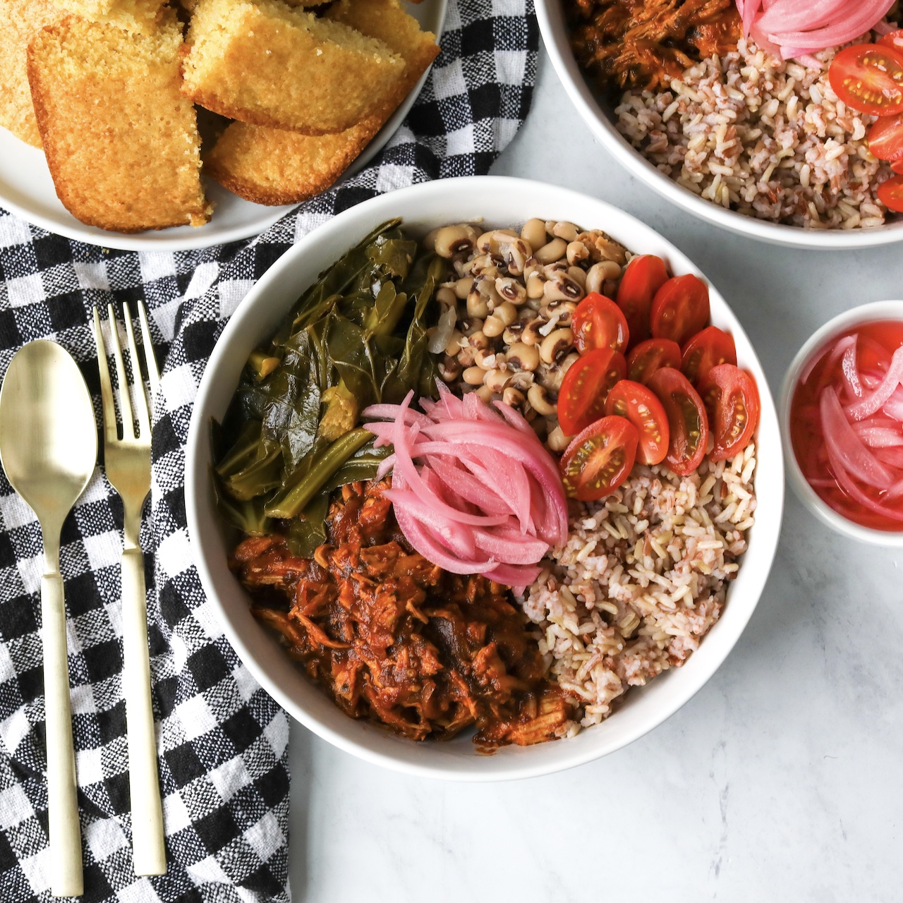 Authentic Soul Food Bowl with BBQ Chicken with onee bowl in focus angled from the top or above the image. Small corner of plated cornbread shown, silverware with fork and spoon, black and white decorative towel for styling purposes and a small bowl of pickled red onion.