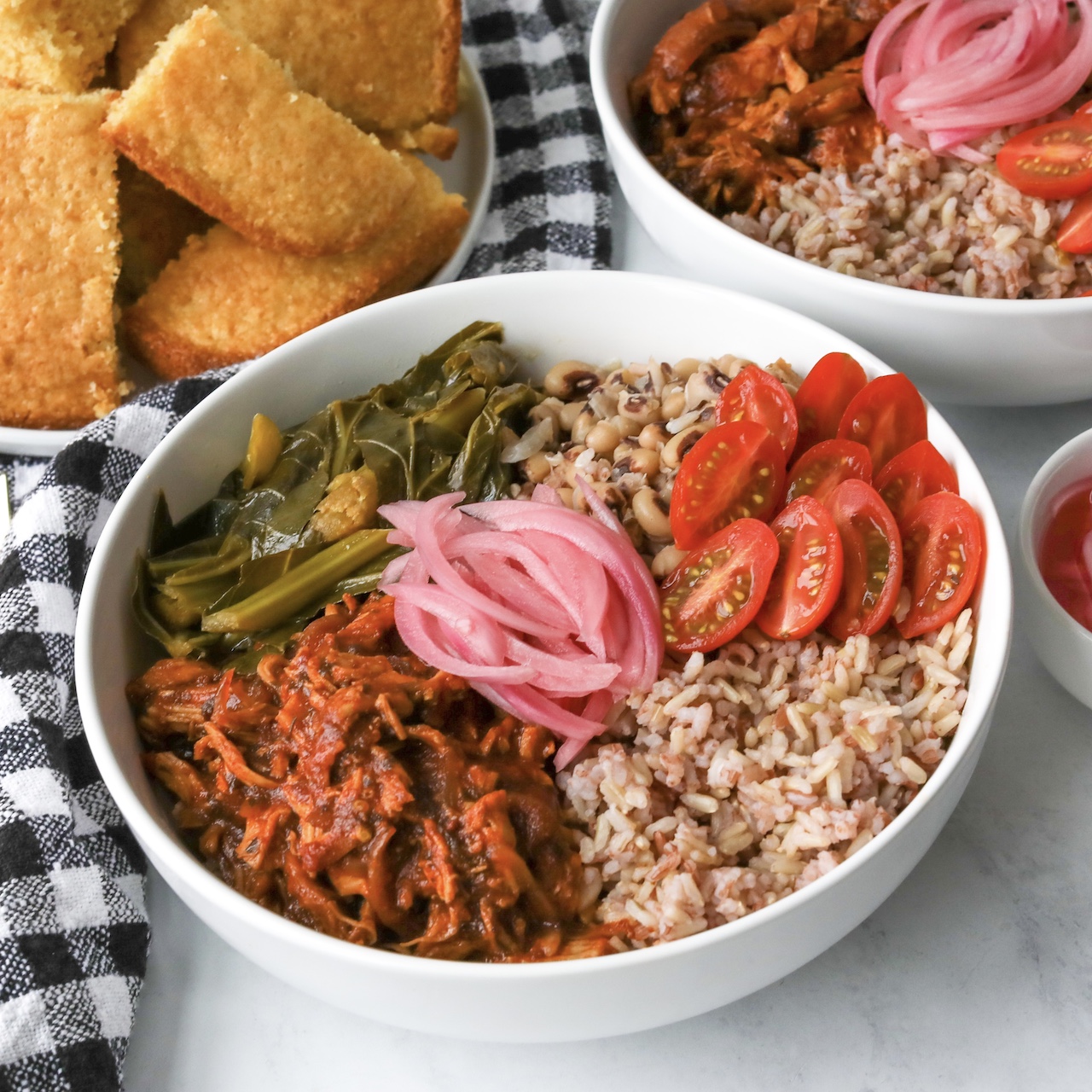 One full bowl and half of a second bowl in frame of Authentic Soul Food Bowl with BBQ Chicken. Plated cornbread on the left side of bowl and additional bbq bowl plate on the right. Black and white decorative towel added for extra styling purposes.