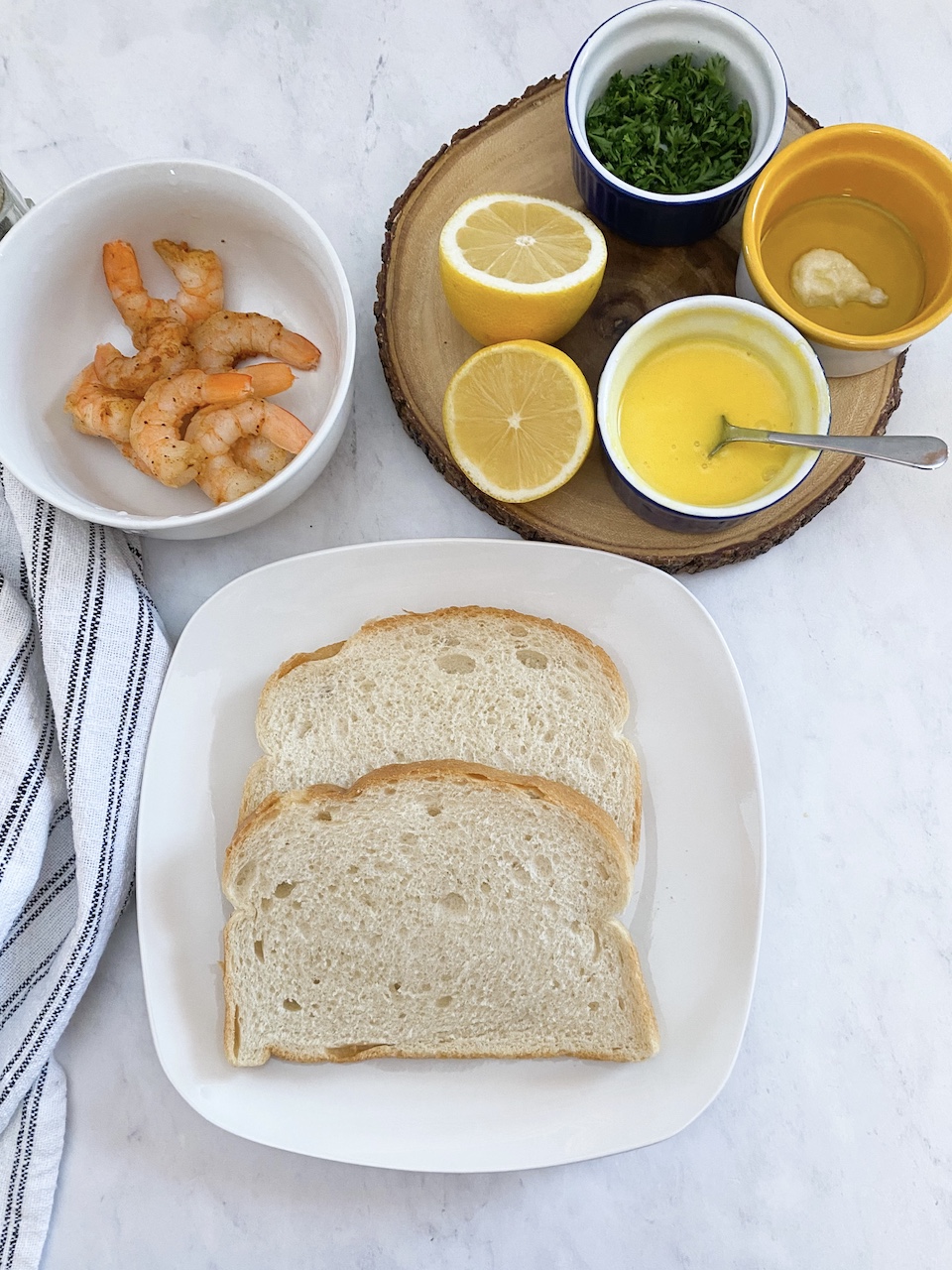 Picture of ingredients; white plate with toast, cajun shrimp in a bowl, lemons sliced, hollandaise sauce in a cup, olive oil and garlic in a bowl and parsley and chives in a small blue bowl on top of a round wooden cutting board