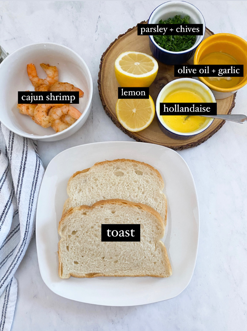 Picture of ingredients with labels; white plate with toast, cajun shrimp in a bowl, lemons sliced, hollandaise sauce in a cup, olive oil and garlic in a bowl and parsley and chives in a small blue bowl on top of a round wooden cutting board