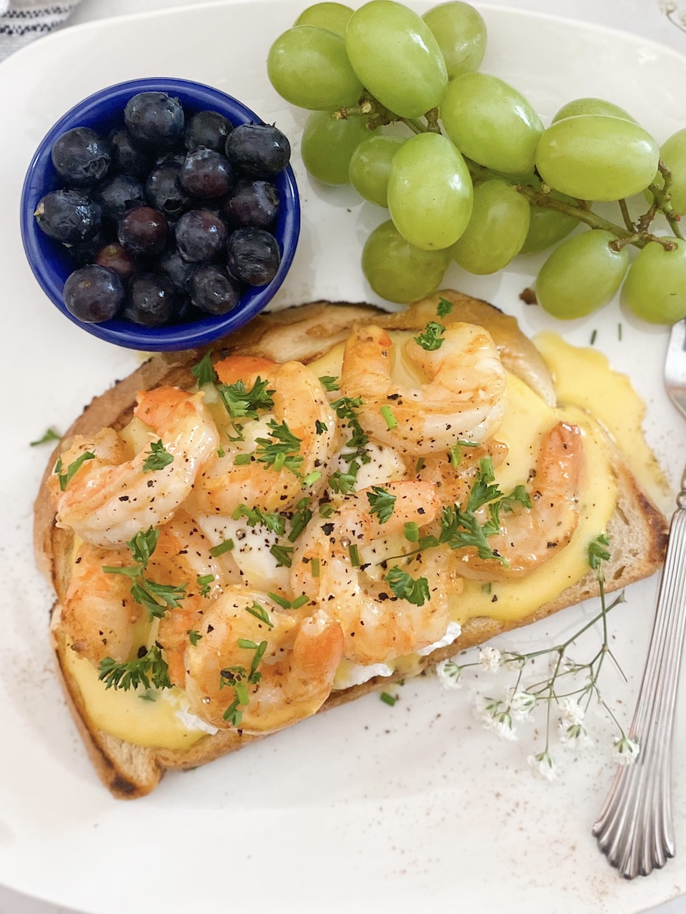 White plate with cajun shrimp toast sliced down the middle to show runny egg and hollandaise sauce. Blueberries, grapes on the plate. Plate is also dressed with babies breath for garnish.