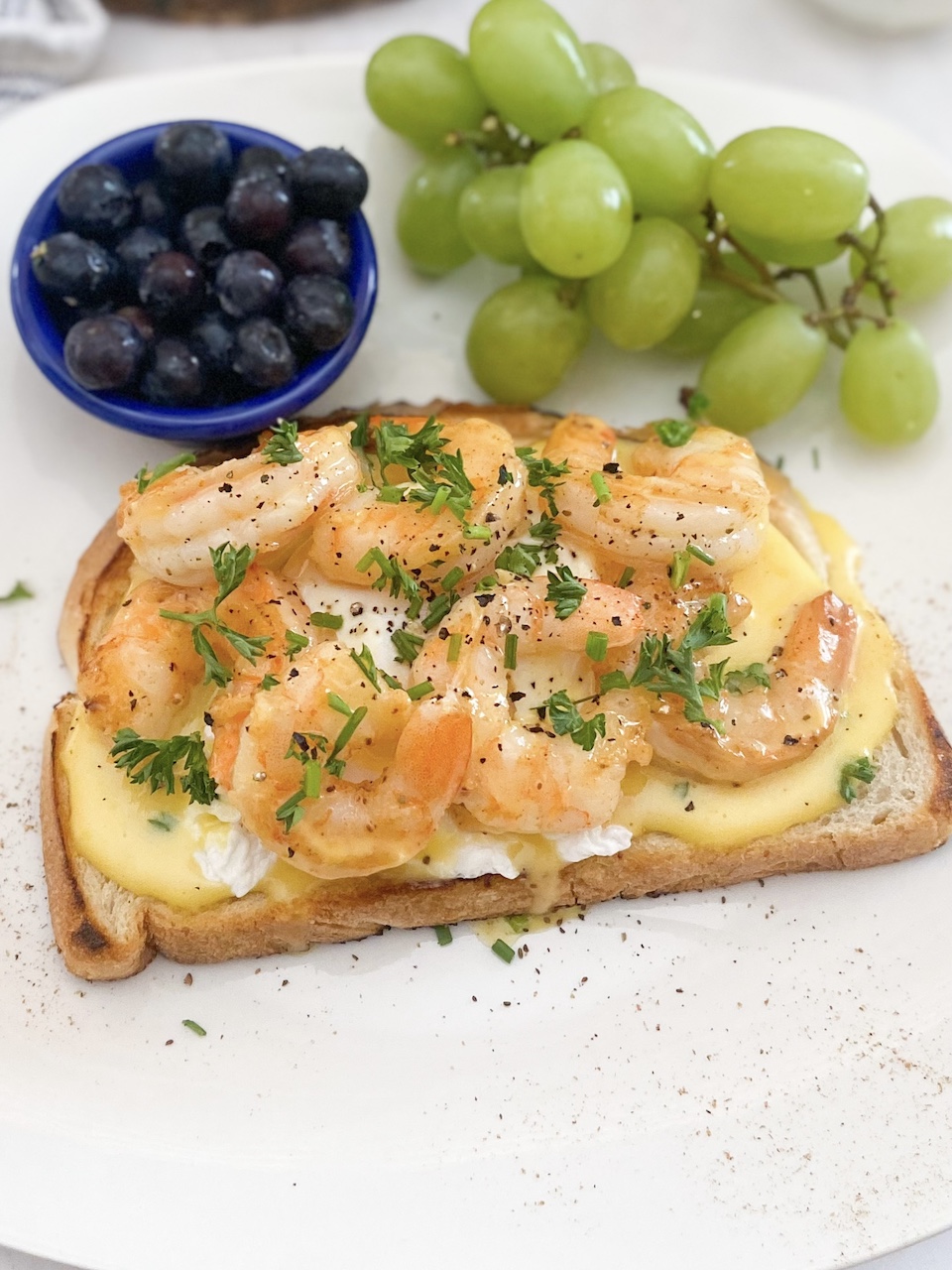 White plate with cajun shrimp toast and hollandaise sauce. Blueberries, grapes on the plate. Plate is also dressed with babies breath for garnish.