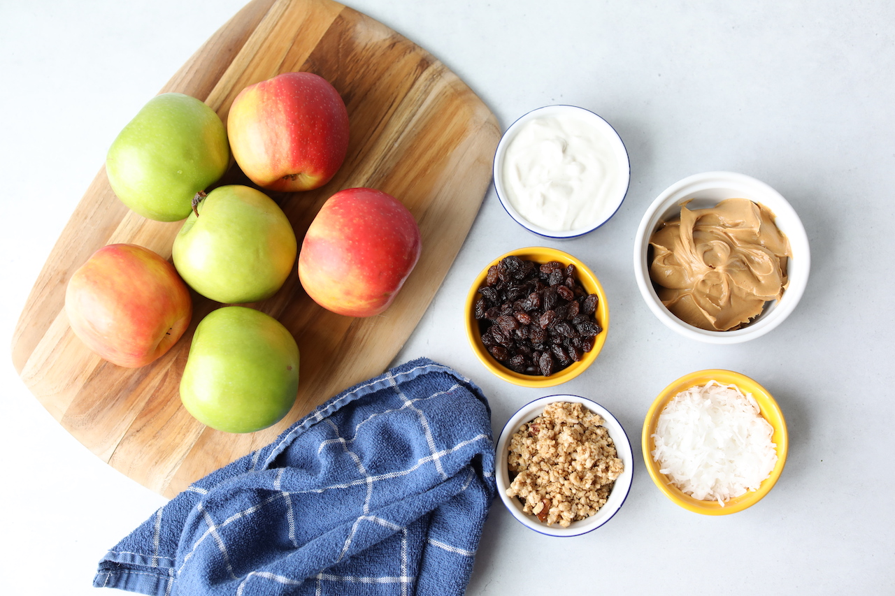 Ingredient lists for apple nachos. Five apples on a cutting board with a decorative blue towel. 5 ramekins filled with toppings for apple nachos.  