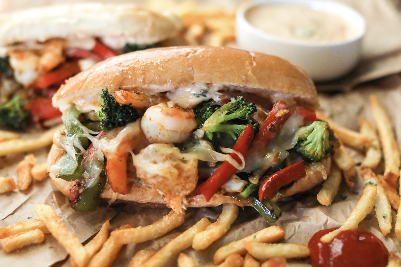 Seafood Cheesesteak centered with red, green peppers, onions and broccoli topped with remoulade sauce. Styled with french fries and ketchup around hoagie with remoulade sauce in white cup not in focus.