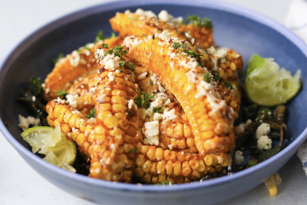 Tik Tok Trend Corn Ribs piled onto a blue bowl with one corn "rib" in focus. Styled with sliced poblano peppers on the bottom and a two wedges of lime.