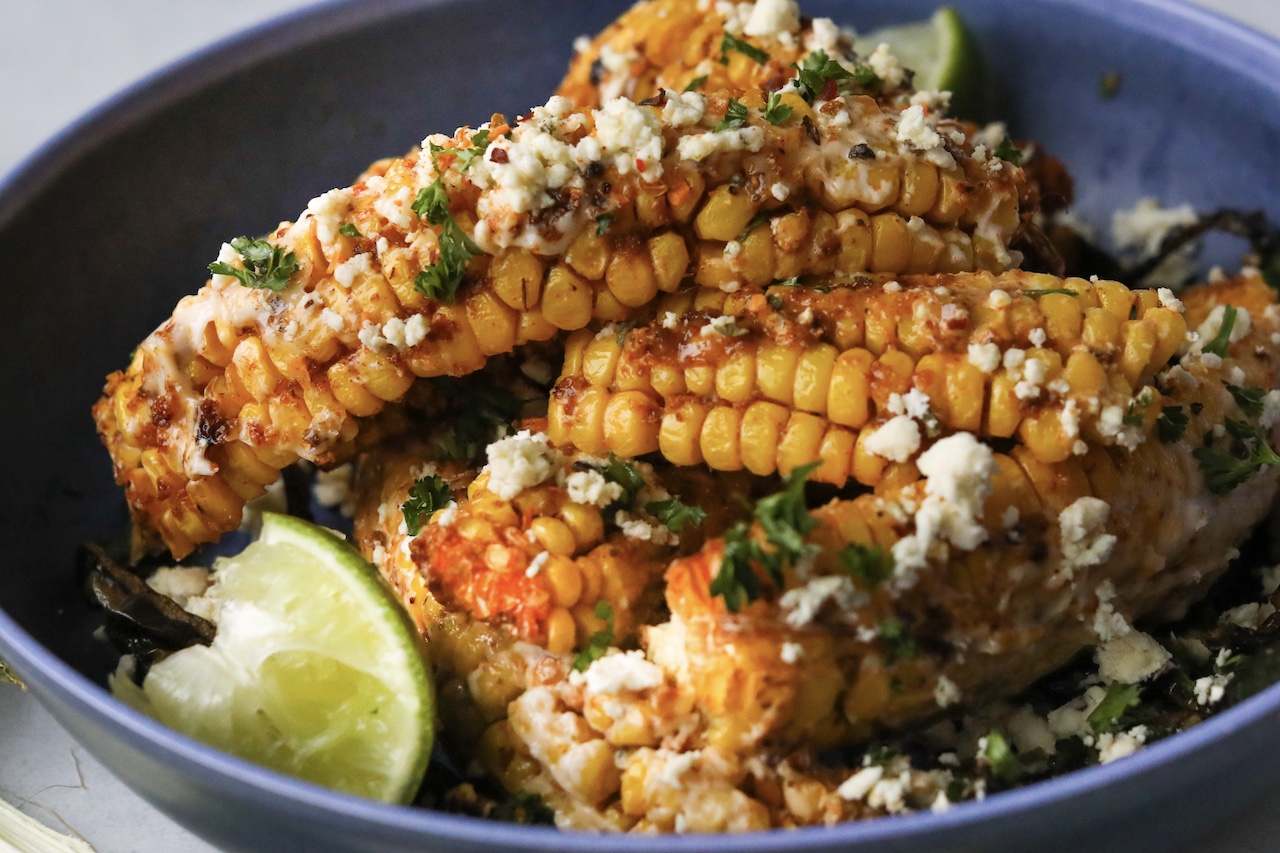 Tik Tok Trend Corn Ribs piled onto a blue bowl with one corn "rib" in focus. Styled with sliced poblano peppers on the bottom and a squeezed wedge of lime.