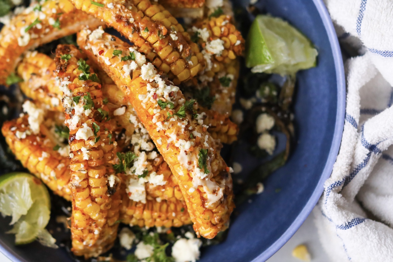 Tik Tok Trend Corn Ribs piled onto a blue bowl with corner of bowl with corn riblets in focus. Styled with sliced poblano peppers on the bottom and a two wedges of lime.