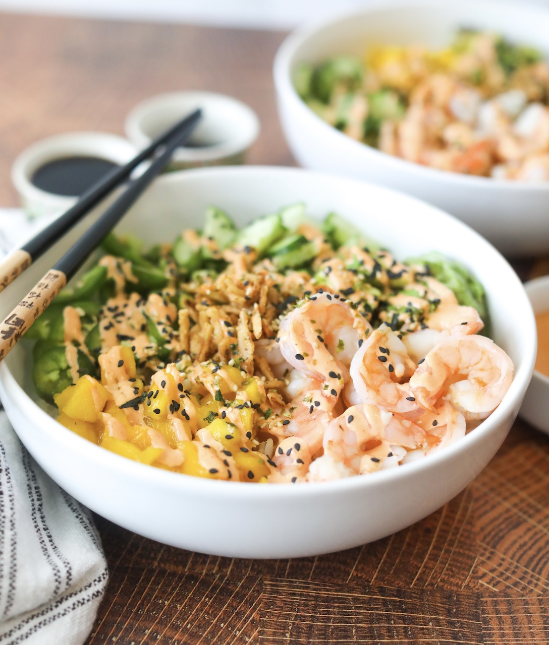 Two bowls with one in focus. Bowl is filled with dragon roll sushi bowl ingredients. Fresh poached shrimp, cucumber, jalapeno, avocado, crunchy onion and mango. Topped with black sesame seeds and furikake. Black and brown vintage chopsticks are laid on top of the left side of the bowl. Two small grey asian hand painted teach cups on the left side out of focus. The left corner of the image has a black and white towel.