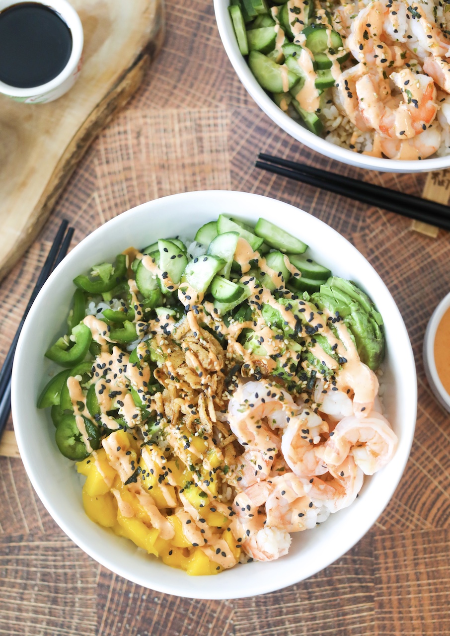 Dragon Roll Sushi Bowl plated in a white howl with ingredients separated around the bowl. Fresh jalapeno, mango, cucumber, shrimp and avocado on top of white sushi rice. Drizzled with spicy mayo and unami sauce and topped with black sesame seeds. 