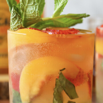 One short glass with strawberry, peach, mint and basil bourbon mash. Drink is topped with fresh basil and additional bottle of honey and another drink not in view in the background.