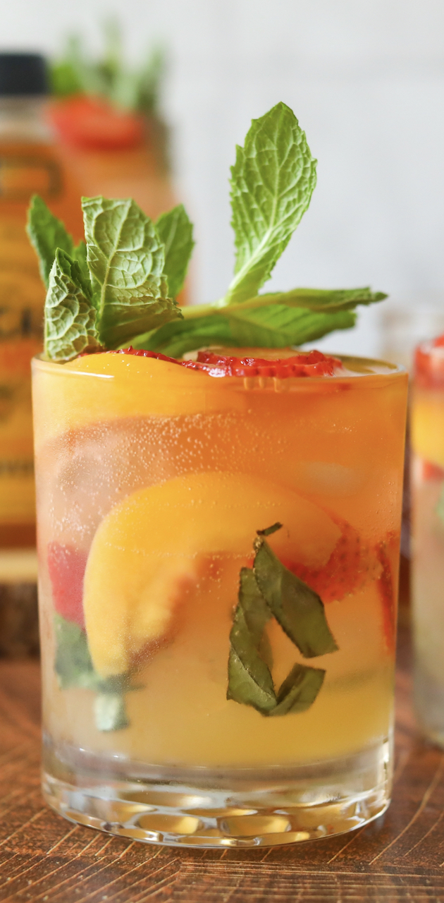 One short glass with strawberry, peach, mint and basil bourbon mash. Drink is topped with fresh basil and additional bottle of honey and another drink not in view in the background.
