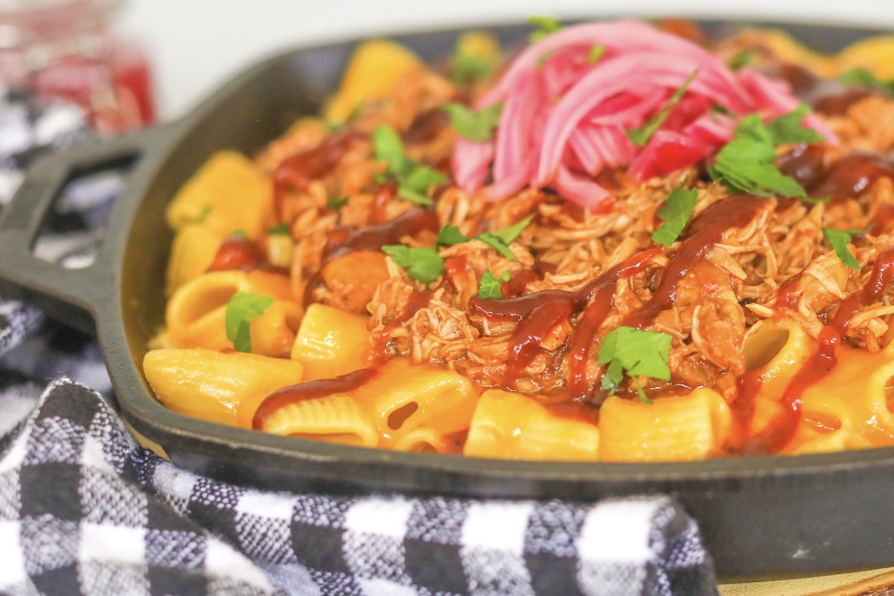 Black cast iron grill pan filled with three cheese mac and cheese topped with shredded bbq chicken and pickled red onions. Black and white towel on the left side of the pan added for style.