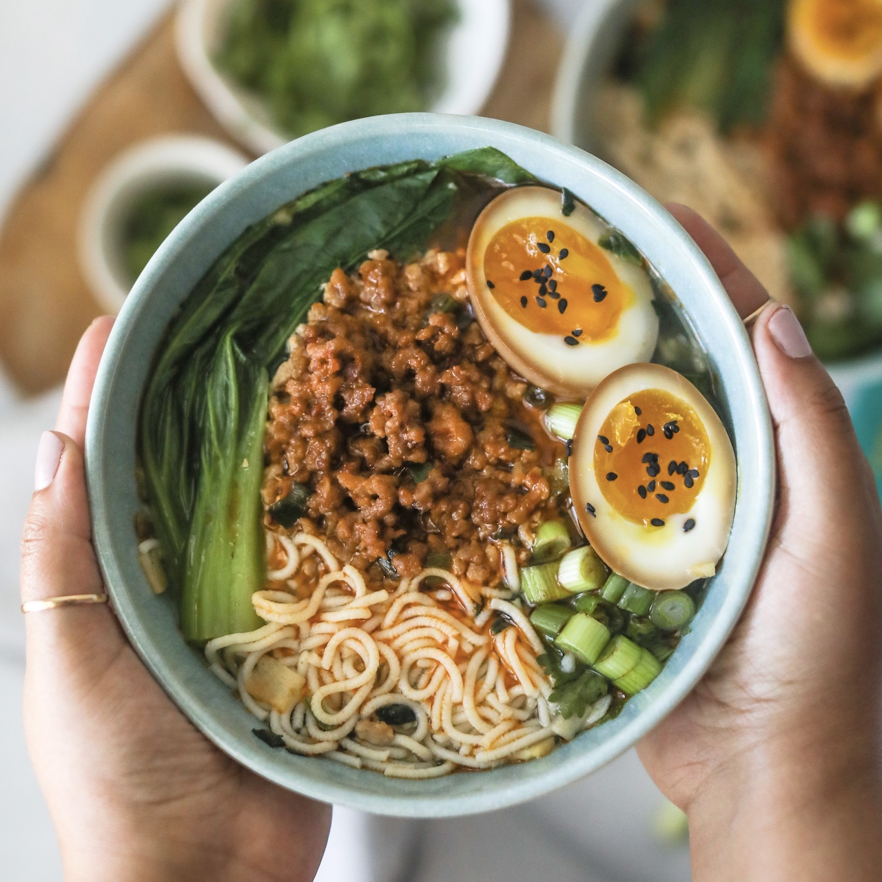 Blue bowl held by two hands with garlic noodle soup cup with spicy ground pork. Out of focus is another blue bowl with recipe and small white bowls with extra toppings for styling purposes.
