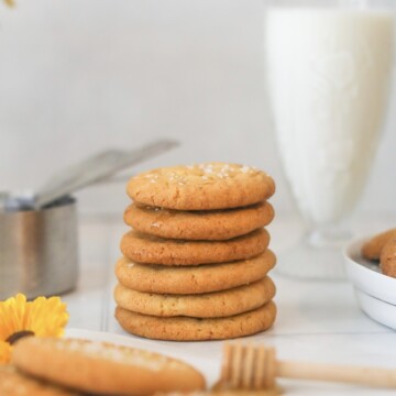 Six stacked honey truffle salted cookies in focus. Out of focus are measuring cups on the left, two cookies in front of the cookie stack, a wooden honey dipper, plated cookies and a glass of milk.