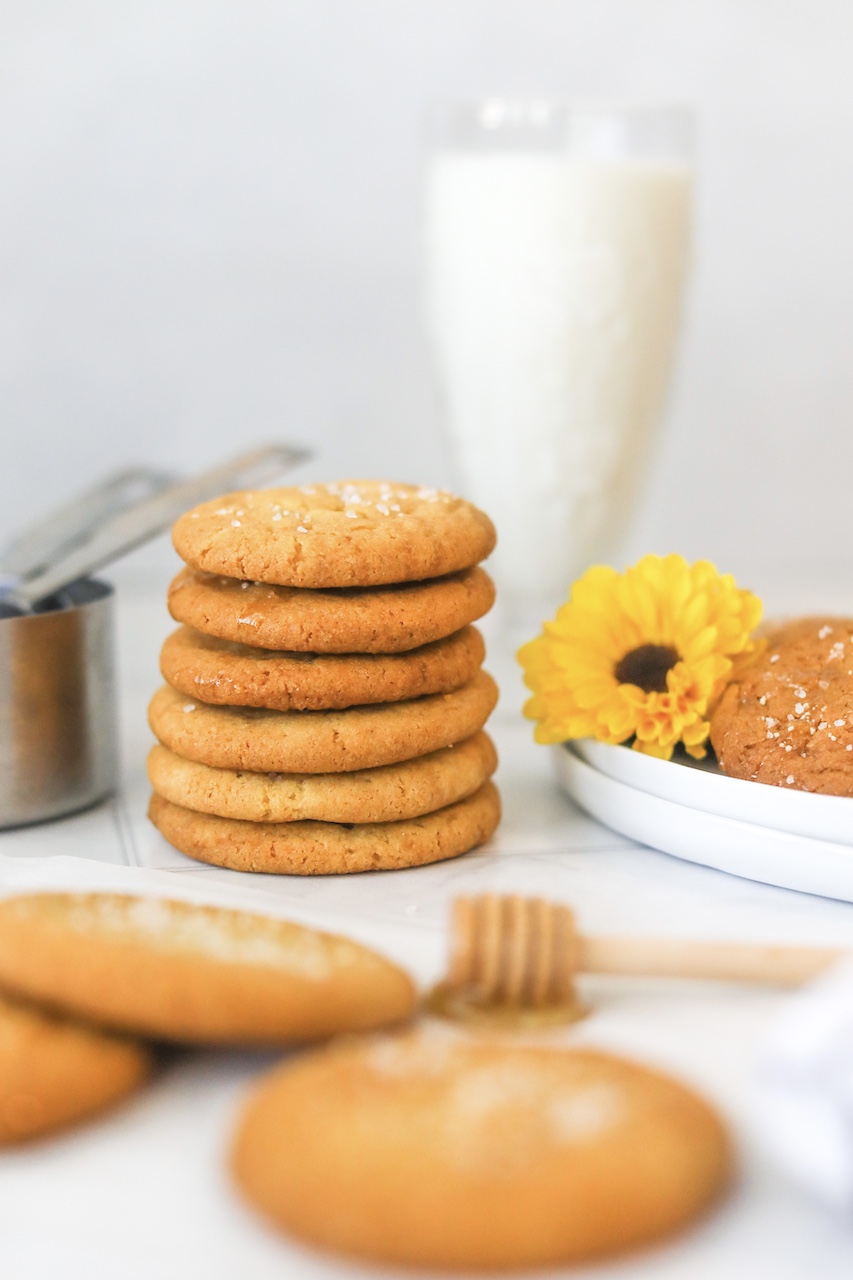 Stack of 6-7 cookies in focus with three flat cookies around the stacked cookies out of focus. Glass of milk behind the stacked cookies for styling purposes. A plate of cookies with a sunflower for added styling and measuring cups on the left side of the cookies for styling purposes.