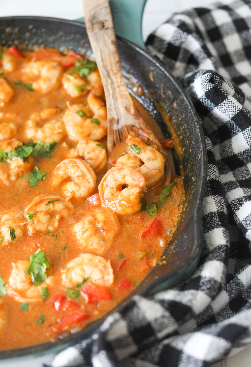 Teal cast iron skillet filled with quick Thai Coconut Shrimp. Black and white checkered kitchen towel added for aesthetics and wooden spoon scooping the shrimp for clearer visibility.