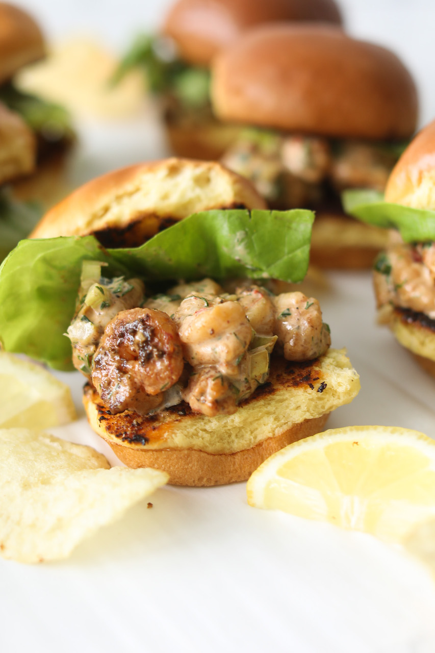 New England Mini Shrimp Rolls close up with otp bun off of roll. Shrimp filling is exposed and green bibb lettuce surrounded by sporadic plain salted chips.