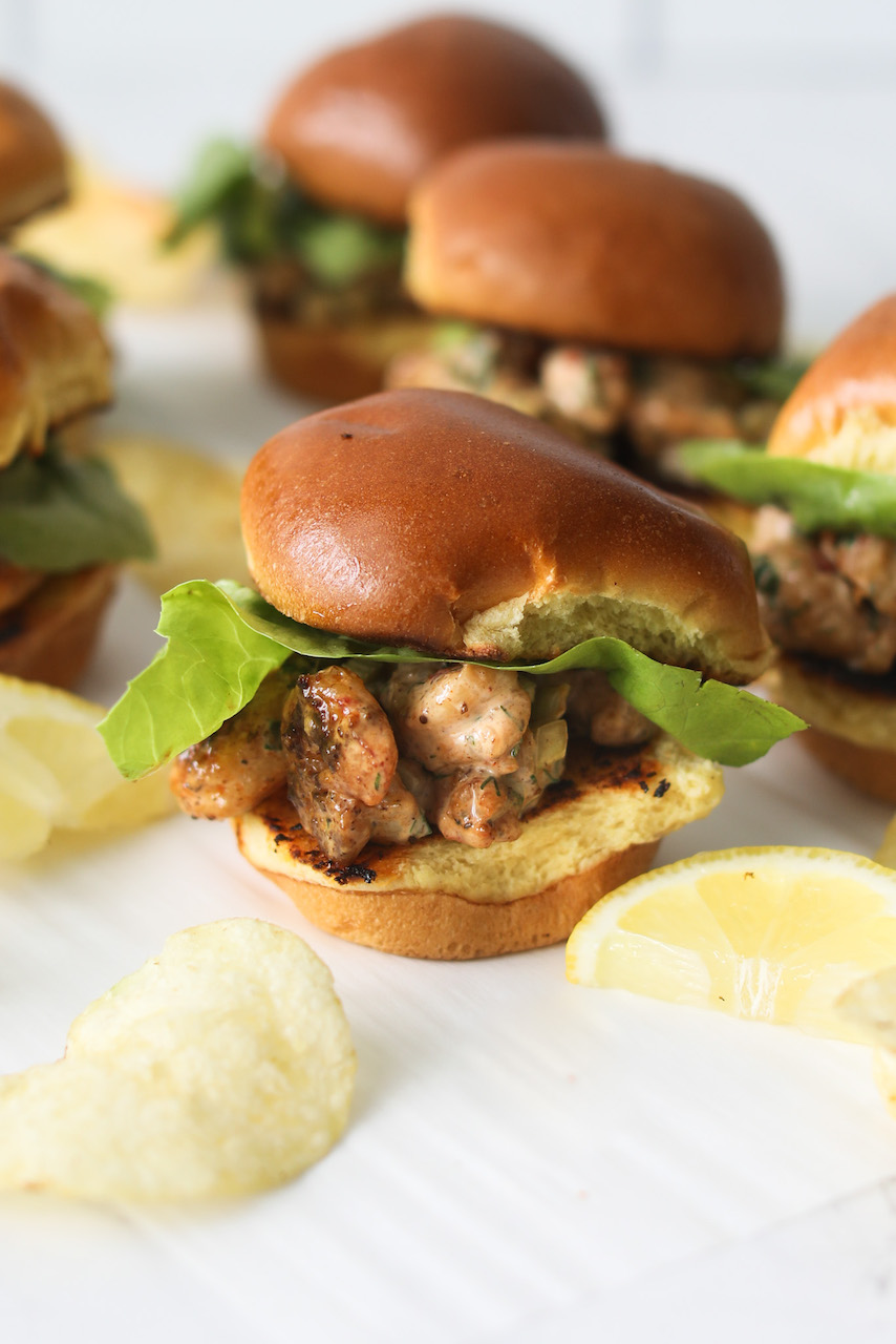 New England Mini Shrimp Rolls close up with otp bun off of roll. One Mini Shrimp Roll is in focus and surrounded by sporadic plain salted chips.