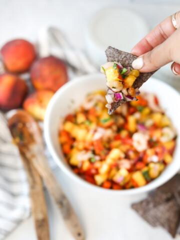Right hand holding blue tortilla chip with peach salsa with shrimp. Out of focus are two wooden spoons, four peaches, white bowl with remaining peach salsa and black and white dish towel used for styling.