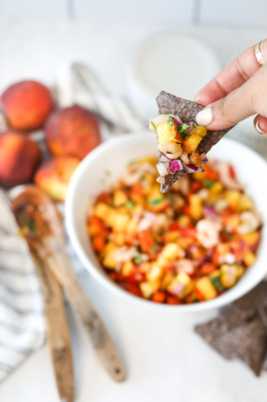 Right hand holding blue tortilla chip with peach salsa with shrimp. Out of focus are two wooden spoons, four peaches, white bowl with remaining peach salsa and black and white dish towel used for styling.