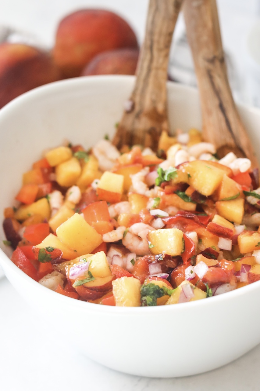 White bowl with peach salsa with shrimp. Three peaches on the left side of the bowl with two wooden spoons and a decorative black and white towel for styling.
