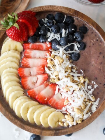 Very Berry Energy Smoothie Bowl poured into a small wooden bowl with wooden spoon on the left side. Filled with purple smoothie and topped with fresh strawberries, bananas, shaved coconut, blueberries and granola. Small clear bowls around the smoothie bowl to style filled with whole strawberries, blueberries and granola.