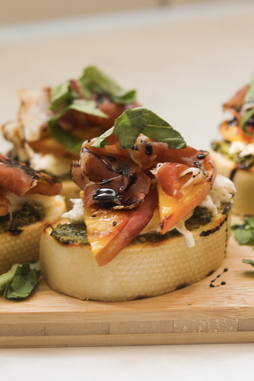 Peach, Prosciutto and Burrata Crostini in focus with three out of focus around. Crostini's are placed on a wooden cutting board with a white background.