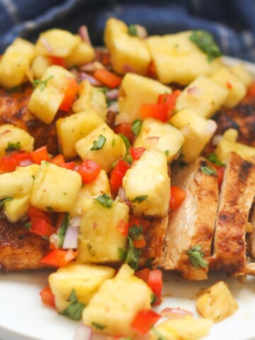 Two white small dinner plates stacked with two chicken grilled blackened chicken breasts with pineapple salsa on top. Out of focus and to the right are two gold forks, blue dish towel for styling purposes.