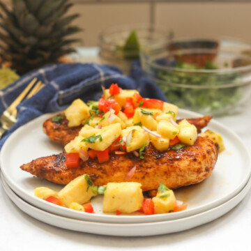 Two white small dinner plates stacked with two chicken grilled blackened chicken breasts with pineapple salsa on top. Out of focus and to the right are two gold forks, blue dish towel for styling purposes.