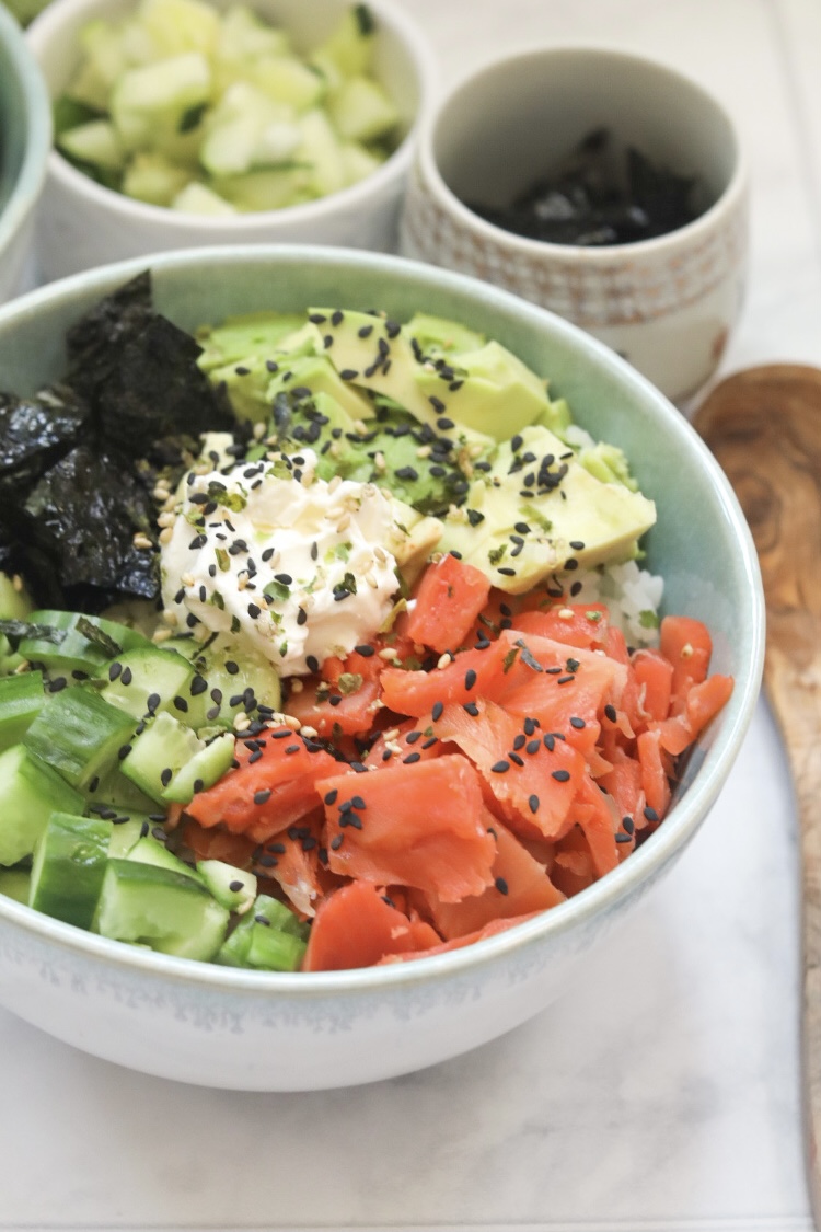 Light blue bowl filled with smoked salmon, cucumber, cream cheese and seaweed snack papers. Dish is topped with black sesame seeds and small bowls in white are surrounding the dish filled with soy sauce and black sesame seeds for styling purposes.