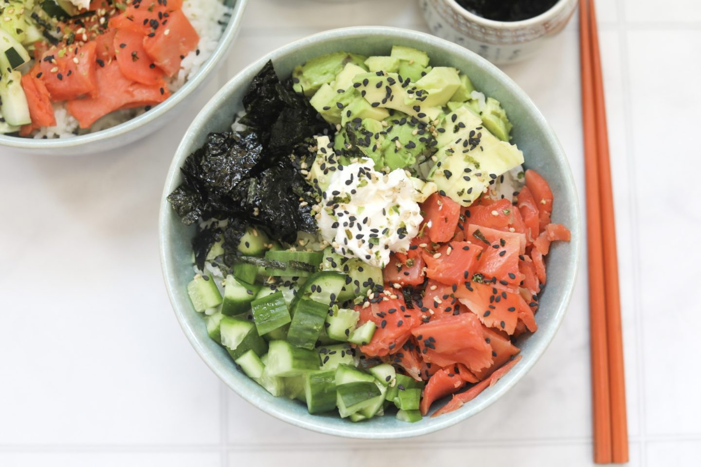 2 Philly Roll Sushi Bowls filled with smoked salmon, cream cheese, avocado, cucumber and nori sheets. Tan colored chopsticks on the right side of the bowl and a small grey bowl filled with soy sauce for styling purporses. 