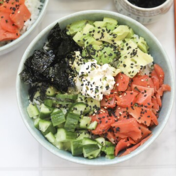 2 Philly Roll Sushi Bowls filled with smoked salmon, cream cheese, avocado, cucumber and nori sheets. Tan colored chopsticks on the right side of the bowl and a small grey bowl filled with soy sauce for styling purporses.