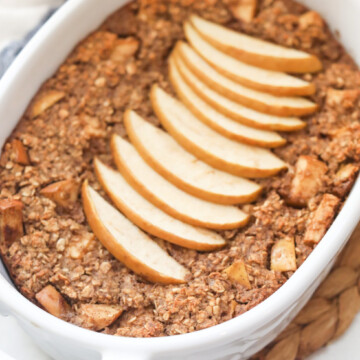 White oval baking dish with baked apple pie oatmeal topped with thin sliced red apples feathered on top. Two apples are out of focus in the top left corner of image.